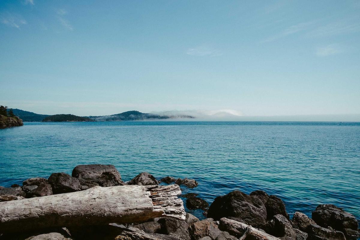 A calm sea with rocky shoreline and distant misty hills under a clear sky.